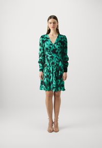 Robe verte avec un motif abstrait noir et turquoise, décolleté en V, manches longues, taille cintrée et ourlet fluide. Finition en tissu lisse.