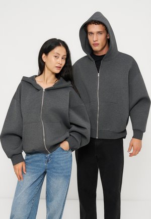 Ung kvinde og mand iført matchende mørkegrå hættetrøjer med lynlås, kvinden med løst hår og jeans, manden med hætten oppe og sorte bukser.