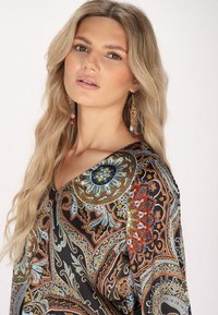 Blusa nera con motivo paisley, caratterizzata da un ornato scollo rotondo e design multicolori intricati. Il modello indossa lunghe orecchini colorati.