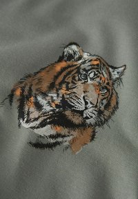 Pelēks audums ar detalizētu izšūtu tīģera galvu oranžā un melnā krāsā ar izsmalcinātām ēnošanas detaļām kažokā un sejas vaibstos.