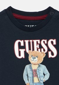 Μπλε ναυτικό μπλουζάκι με στρογγυλή λαιμόκοψη και μπορντό εσωτερικό γιακά, με γραφικό αρκούδας που φοράει μπουφάν και μπλουζάκι και το κείμενο "GUESS" με έντονα γράμματα.