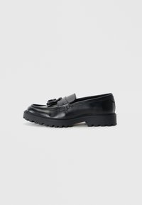 Mocasines de cuero negro con un acabado brillante, suela de goma gruesa y detalle decorativo de borlas en la parte delantera.