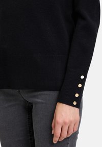 Schwarzer Strickpullover mit geripptem Saum und Bündchen. Die Bündchen sind mit vier goldfarbenen Knopfakzenten versehen. Lange Ärmel, weiche Textur, figurbetontes Design.