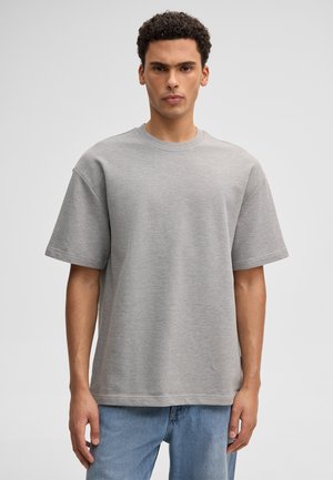 Homme aux cheveux courts et bouclés portant une chemise grise unie à manches courtes surdimensionnée et un jean bleu, de face sur un fond uni.