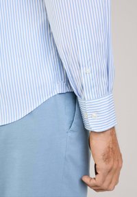 Camisa de rayas azul claro y blanca de manga larga con puños abotonados, que presenta un dobladillo curvado. Tejido de algodón, textura suave.