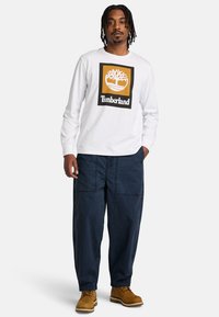 Camicia bianca a maniche lunghe con un logo Timberland in un quadrato nero e oro, abbinata a pantaloni cargo blu scuro e larghi e stivali color sabbia.