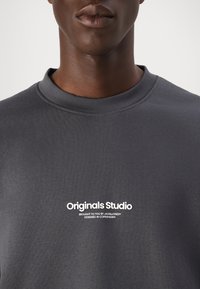 Sivá mikina s rebrovaným okrúhlym výstrihom, s bielym nápisom "Originals Studio" a ďalším malým textom. Hladká, jemná textúra látky.