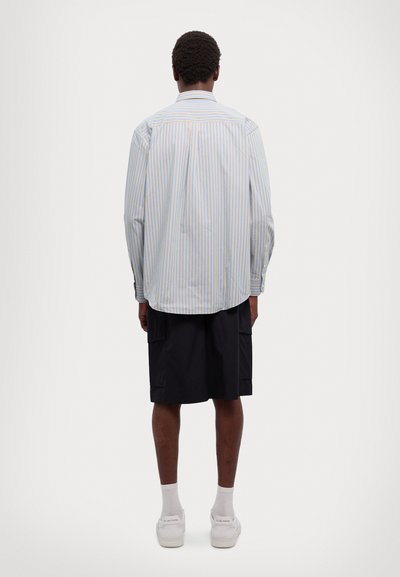 Camicia con bottoni, a righe verticali blu e gialle, taglio aderente, maniche lunghe e orlo curvo; abbinata a pantaloni cargo neri.