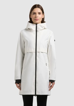 Woman wearing a white hooded long coat with black zipper and black cuffs, standing against a plain light background.