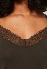 Olivgrön linne med V-ringning och mörk spetsdetaljering samt fransar längs kanterna. Mjuk, slät tygtextur.