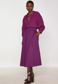 Donna che indossa una blusa lunga manica viola, una gonna midi a vita alta abbinata, stivali con tacco marroni e orecchini a cerchio dorati, in piedi contro uno sfondo bianco.
