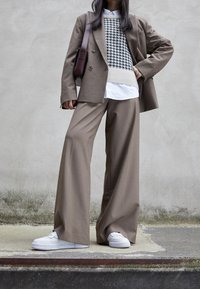 Veste de costume marron sur un pull à motif pied-de-poule et une chemise blanche, associée à un pantalon à larges jambes et des baskets blanches.