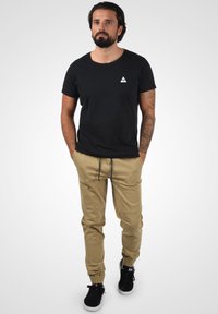 Svart bomull T-skjorte med en liten hvit logo, kombinert med beige joggebukser med elastiske mansjetter og snøring i livet. Svarte sneakers.