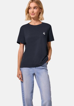 Femme blonde portant un t-shirt noir uni avec un petit logo cerise blanc et un jean bleu clair, debout avec une main dans la poche.