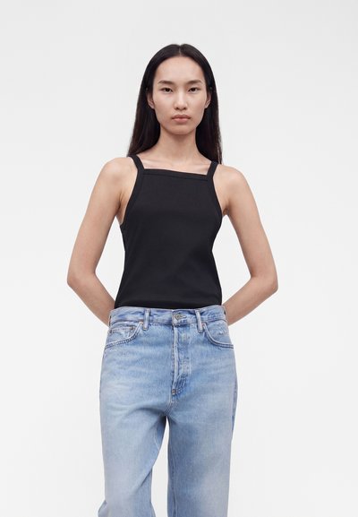 Νέα γυναίκα με μακριά σκουρόχρωμα μαλλιά που φοράει μαύρο αμάνικο τοπ με τετράγωνο ντεκολτέ και ανοιχτό μπλε ψηλόμεσο τζιν, στέκεται με τα χέρια πίσω από την πλάτη.