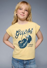 Dzeltena kokvilnas T-kreklā ar sarullētām piedurknēm, ar tumši zilu grafiku, kurā attēlotas ķirši, un tekstu "Guess est. 1981 California Los Angeles".