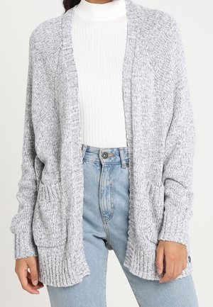 Cardigan en maille épaisse gris clair avec poches devant, porté sur un col roulé côtelé blanc et un jean taille haute bleu clair.