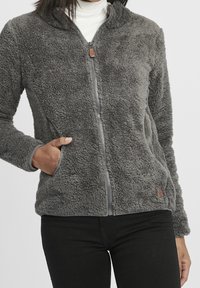 Harmaa fleece-takki, jossa on täysi etuvetoketju, kaksi kylkitaskua ja teksturoitu pinta. Takissa on kontrastivärinen reunus ja pieni logopatch.