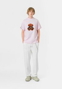 Camiseta de manga corta rosa claro con un gráfico de osito de peluche, combinada con pantalones blancos y zapatillas verdes de tipo slip-on. Estilo casual, ajuste relajado.