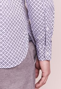 Svetle fialová košile s geometrickým černým vzorem, s knoflíkovým manžetovým lemem a zakřiveným spodním lemem. Spárována se šedým materiálem níže.