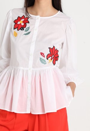 Blouse blanche boutonnée avec broderie florale rouge, taille froncée, manches ballon trois-quarts, associée à un pantalon rouge vif.