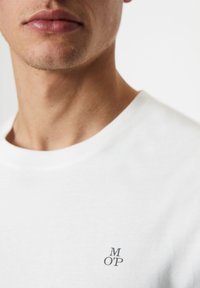 Camiseta de algodón blanca con cuello redondo. Presenta un pequeño logo impreso "M O’P" en la zona izquierda del pecho. Textura suave y diseño casual.