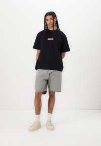 Black t-shirt with pink graphic, paired with light grey shorts. Model wears beige sneakers and white socks. Minimalist background.