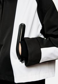 Schwarz-weiße Jacke mit einem eleganten Design, ausgestattet mit einer Reißverschlusstasche am Ärmel und strukturiertem Stoff. Betonte Bündchen und Nähte.