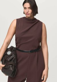Bordeaux mouwloos jurk met gedrapeerde halslijn en zijplooien, zwarte leren riem om de taille, met een zwarte leren tas met ritsdetails.