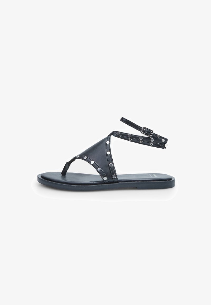 Svart sandal med tårem, ankelsnören och runda metallaccenter. Slätt material, platt sula och minimalistisk design.