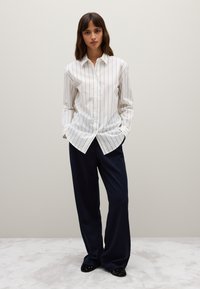 Mujer de pie con las manos en los bolsillos, vistiendo una camisa de rayas blanca de manga larga, pantalones amplios azul marino y zapatos negros sobre una alfombra clara.