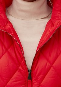 Gilet matelassé rouge avec une finition brillante, doté d'une fermeture éclair sur le devant et d'un col montant, superposé sur un pull à col roulé beige.