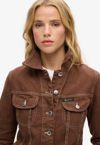 Veste en denim marron avec un col haut, des boutons argentés, deux poches avant et des coutures blanches contrastées. Présente une étiquette de marque.