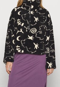 Pullover in pile nero con colletto alto, caratterizzato da motivi astratti bianchi e chiusura con zip frontale. Abbinato a una gonna viola.