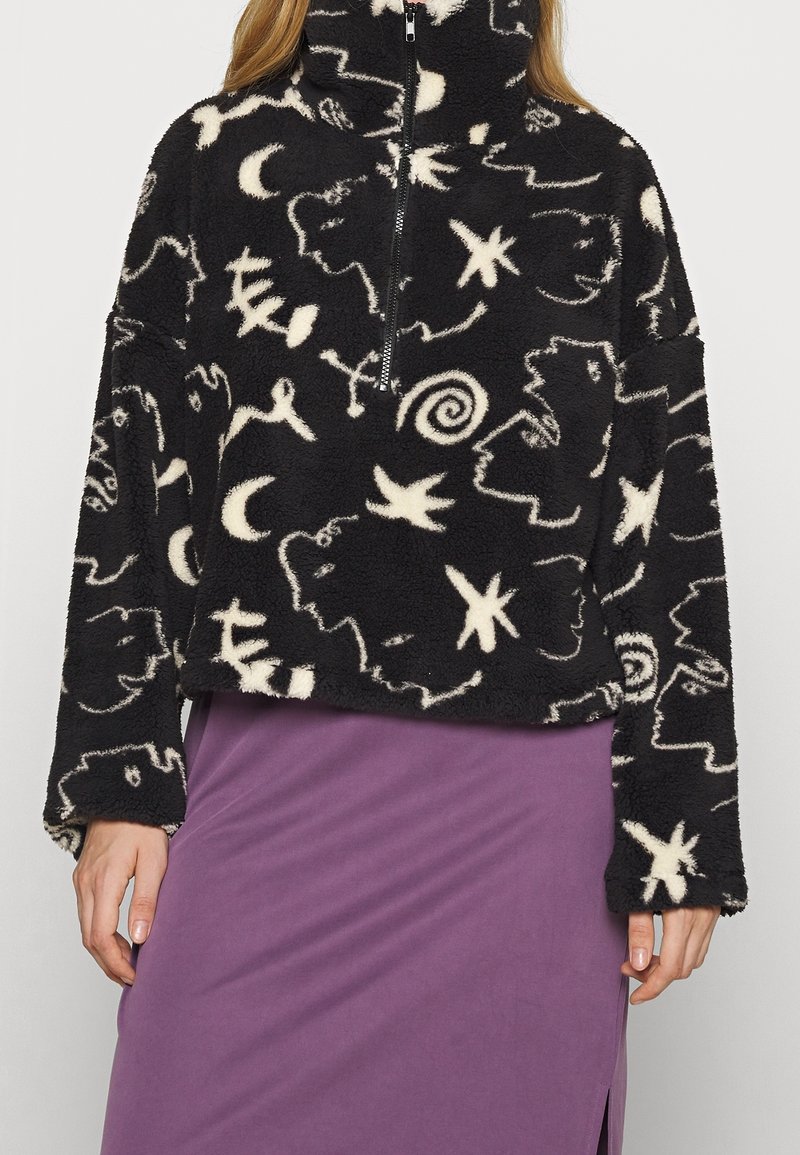Pullover in pile nero con colletto alto, caratterizzato da motivi astratti bianchi e chiusura con zip frontale. Abbinato a una gonna viola.