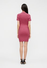 Εφαρμοστό, ριμπ πλεκτό φόρεμα σε σκούρο ροζ με κοντά μανίκια και γιακά πόλο, συνδυασμένο με μαύρα ψηλοτάκουνα παπούτσια. Λεία υφή.