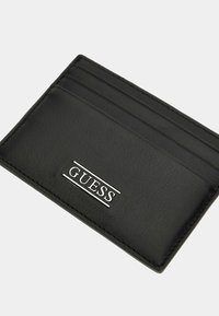 Černý kožený držák karet s čtyřmi přihrádkami, hladkým povrchem a stříbrným logem "GUESS" na přední straně. Kompaktní obdélníkový tvar.