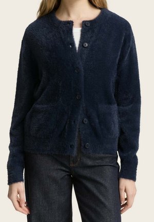 Femme portant un cardigan bleu marine duveteux à boutons avec deux poches avant, par-dessus un haut blanc et un jean foncé.