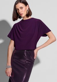 Fioletowy top z krótkimi rękawami z drapowaniem, zestawiony z błyszczącą spódnicą z fioletowych cekinów. Spódnica ma wyjątkowy teksturowany wzór; gładka, śliska tkanina.