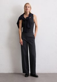 Schwarzes ärmelloses Top kombiniert mit weiten schwarzen Hosen. Ein schwarzer Schal ist über die Schultern gelegt. Glatte Texturen, minimalistisches Design.