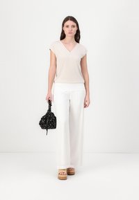 Beige v-hals topp, hvite vide bukser, tresandaler med plattform, holder en svart snølåsveske med blomsterdesign, står mot en nøytral bakgrunn.