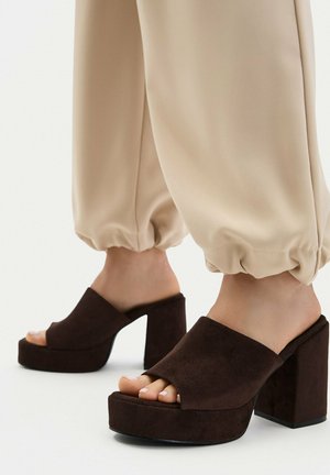 Piedi con mules platform con tacco a blocco in camoscio marrone scuro e punta aperta, indossate con pantaloni beige risvoltati.