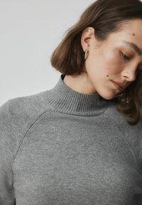 Nuori nainen, jolla on lyhyet ruskeat hiukset, kultaiset renkaankorvakorut ja harmaa ribbineuleinen pooloneule, katsoo alas oikealle.