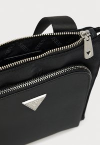 Μαύρη υφασμάτινη τσάντα με ανάγλυφη επιφάνεια, ασημένια φερμουάρ και τριγωνική λεπτομέρεια λογότυπου. Διακρίνονται δύο θήκες με φερμουάρ.