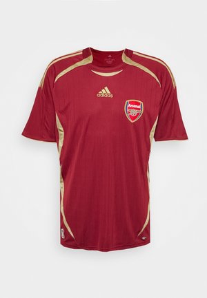 Punainen jalkapallo-paita kultaisilla yksityiskohdilla, pystysuorat raidat, lyhyet hihat ja pyöreä kaula-aukko. Vasemmassa rinnassa on Arsenal-logo.