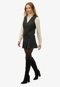 Schwarze Nadelstreifenweste, cremefarbener Rollkragenpullover, plissierter schwarzer Rock und knielange schwarze Stiefel. Slim Fit; strukturierte Stoffe; Knopfdetails an der Weste.