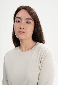 Hellgraues Langarmshirt mit rundem Ausschnitt, das eine glatte Textur und eine lockere Passform aufweist, getragen von einer Person mit glattem braunem Haar.