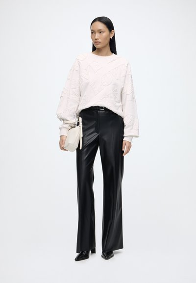 Femme portant un pull blanc avec le texte « LOVE », un pantalon en cuir noir, des chaussures noires et tenant un petit sac à main blanc, debout devant un fond uni.