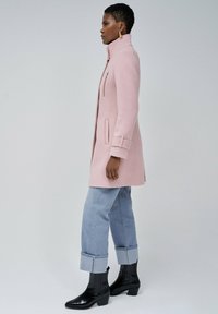 Abrigo de lana rosa claro con cuello alto, bolsillo con cremallera en la parte delantera y mangas con puños. Combinado con unos pantalones vaqueros azules claros remangados y botas negras hasta el tobillo.