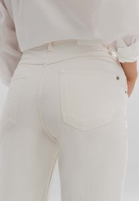 Jeans de denim blanco con una textura suave, que cuentan con dos bolsillos traseros angulados, costuras mínimas y un pequeño detalle de botón de metal.